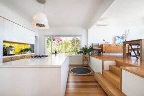 A modern kitchen with a central island, wooden flooring, and a view of greenery outside. - Couples Corner A Stylish Bay View Retreat (McCrae)