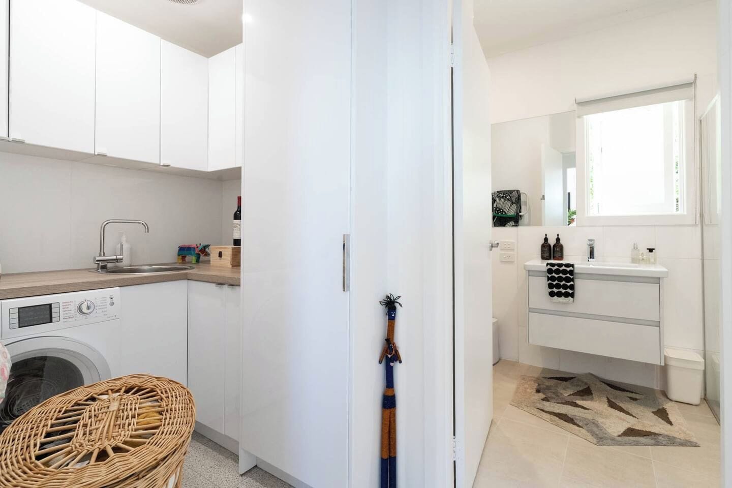 Een compacte keuken met een wasmachine, een spoelbak en een klein eethoekje met een rieten stoel.