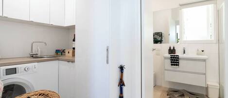 A compact kitchen with a washing machine, a sink, and a small dining area with a wicker chair.