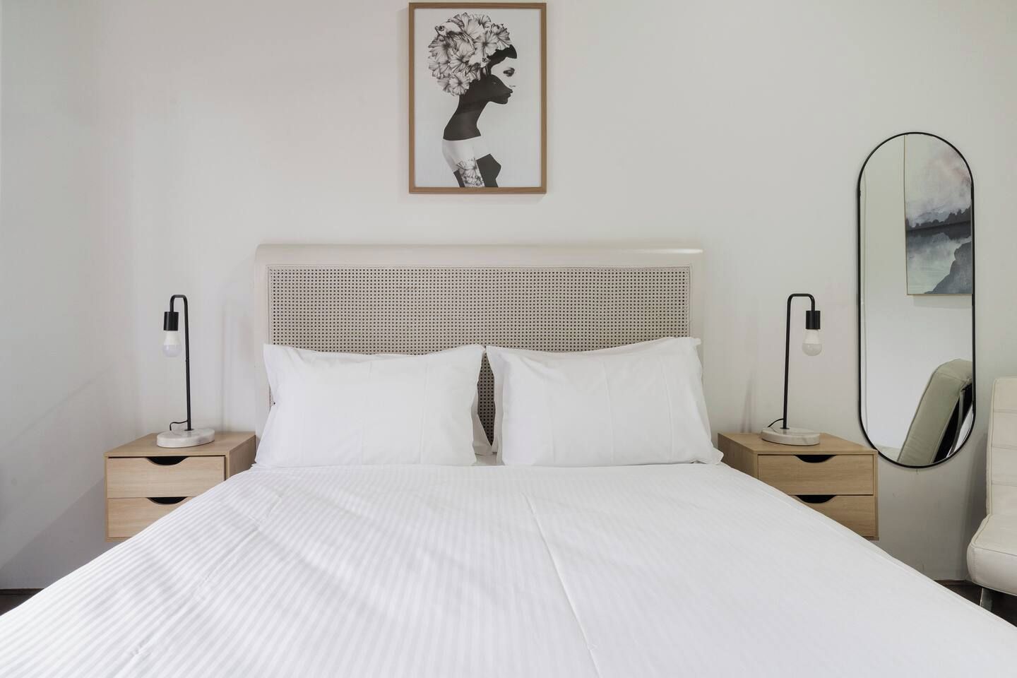 Una cama bien hecha con ropa de cama blanca, dos mesitas de noche con lámparas, un espejo instalado en la pared y una obra de arte enmarcada sobre la cama.