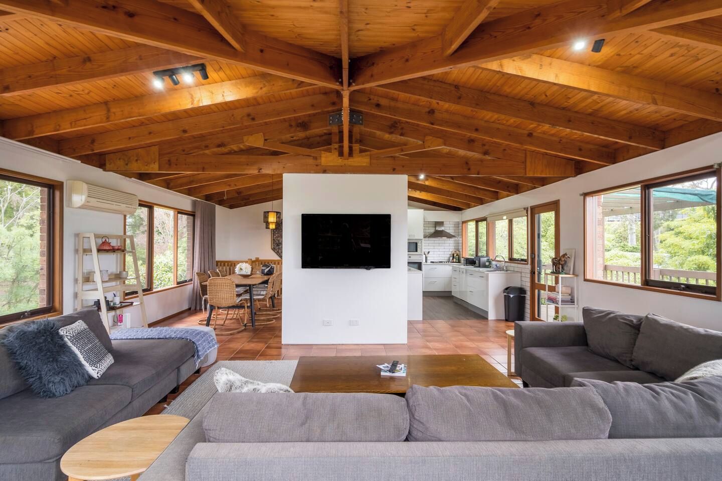 A spacious living area with a high wooden ceiling, a central seating area with sofas, a dining table, and a kitchen in the background.
