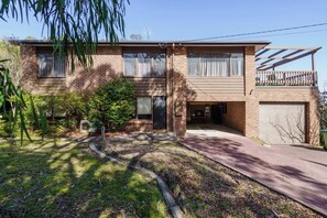 A two-story brick house with a front garage, a covered patio, and a small front yard with a lawn and a tree. - McRae Hillside Terrace Panoramic Family Living (McCrae)