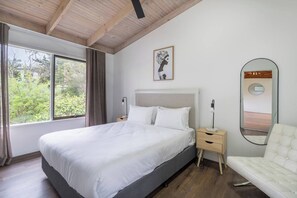 Un dormitorio con una cama grande, mesitas de noche, una silla, un ventanal con cortinas, una obra de arte enmarcada y un espejo.
