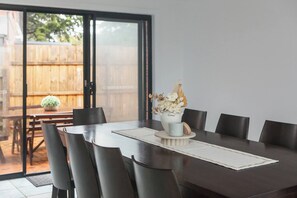 A dining area with a dark wooden table, six chairs, a vase with flowers, and a view of a wooden fence through a glass door. - Hearthstone Haven Spacious Beachside Group Getaway (Safety Beach)