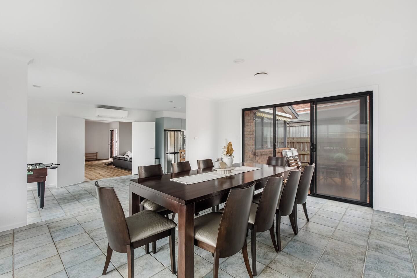 Un comedor moderno con una mesa grande y sillas, una cocina al fondo y un ventanal corredizo que da a un espacio exterior.