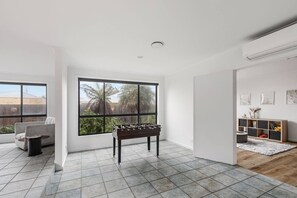 A spacious room with a foosball table, large windows, a sofa, and a wooden cabinet. - Hearthstone Haven Spacious Beachside Group Getaway (Safety Beach)