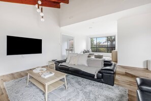 A modern living room with a flat-screen TV, a black leather sofa, a wooden coffee table, and a large window providing a view of the outdoors. - Hearthstone Haven Spacious Beachside Group Getaway (Safety Beach)