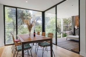 A dining area with a wooden table, wicker chairs, and a bottle of wine. - Ti Tree House Live like a Bonbeach Local (Bonbeach)