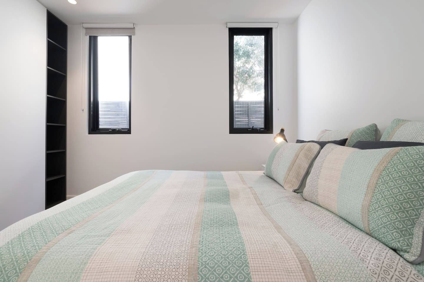 Ein Schlafzimmer mit einem Bett, zwei Fenstern, einem Nachttisch und einer eingebauten Regallösung.