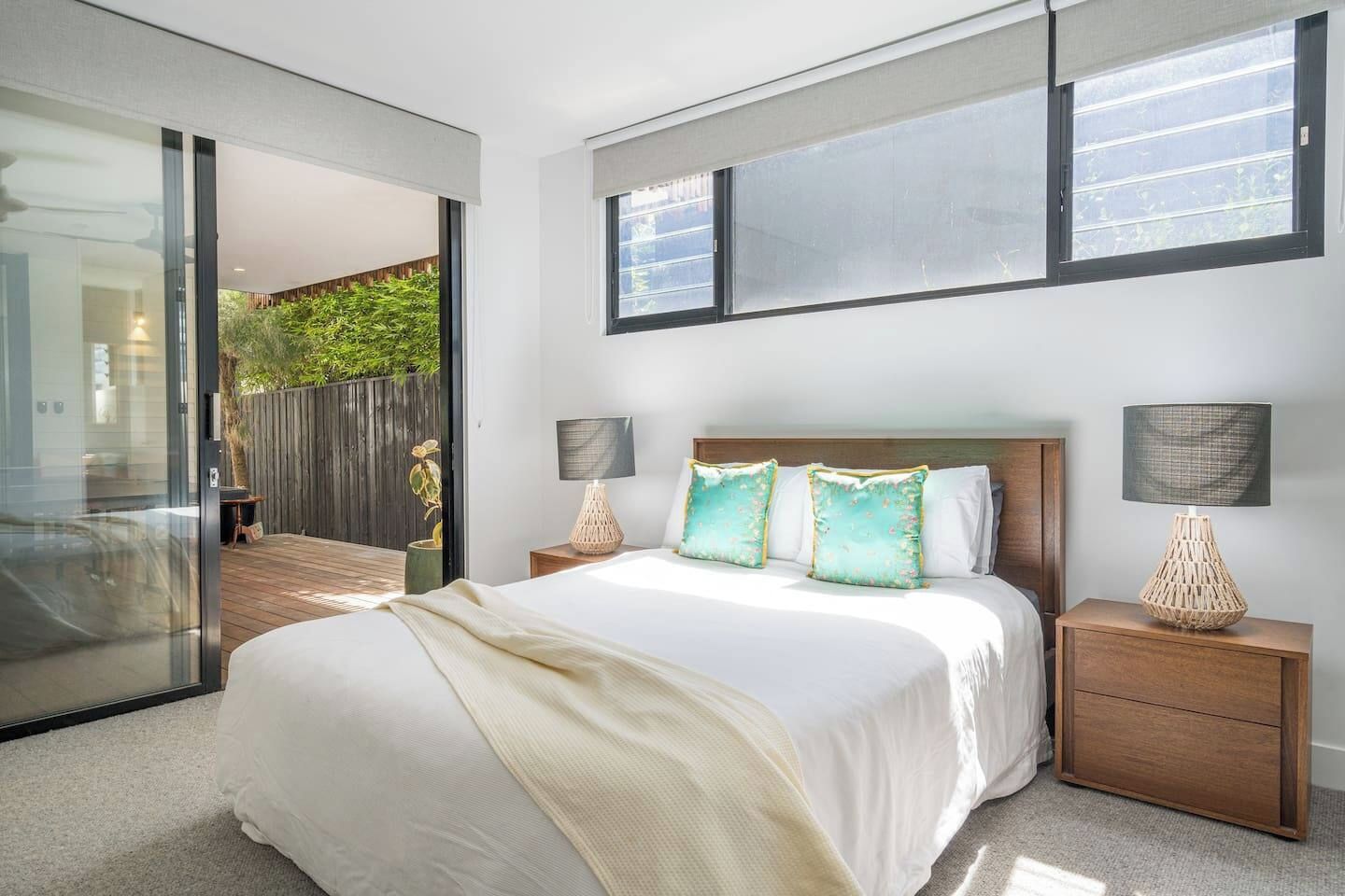 A bedroom with a large bed, two bedside tables with lamps, a sliding glass door leading to a balcony, and a view of greenery outside.