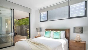Un dormitorio con una cama grande, dos mesitas de noche con lámparas, una puerta corrediza de vidrio que da a un balcón y vistas al exterior con vegetación.
