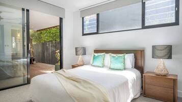 A bedroom with a large bed, two bedside tables with lamps, a sliding glass door leading to a balcony, and a view of greenery outside.