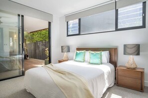 A bedroom with a large bed, two bedside tables with lamps, a sliding glass door leading to a balcony, and a view of greenery outside. - Ti Tree House Live like a Bonbeach Local (Bonbeach)