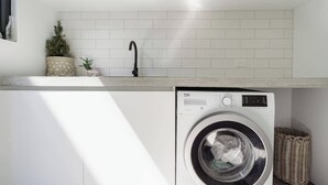 A compact laundry room with a front-loading washing machine, a sink, and a small potted plant. - Ti Tree House Live like a Bonbeach Local (Bonbeach)