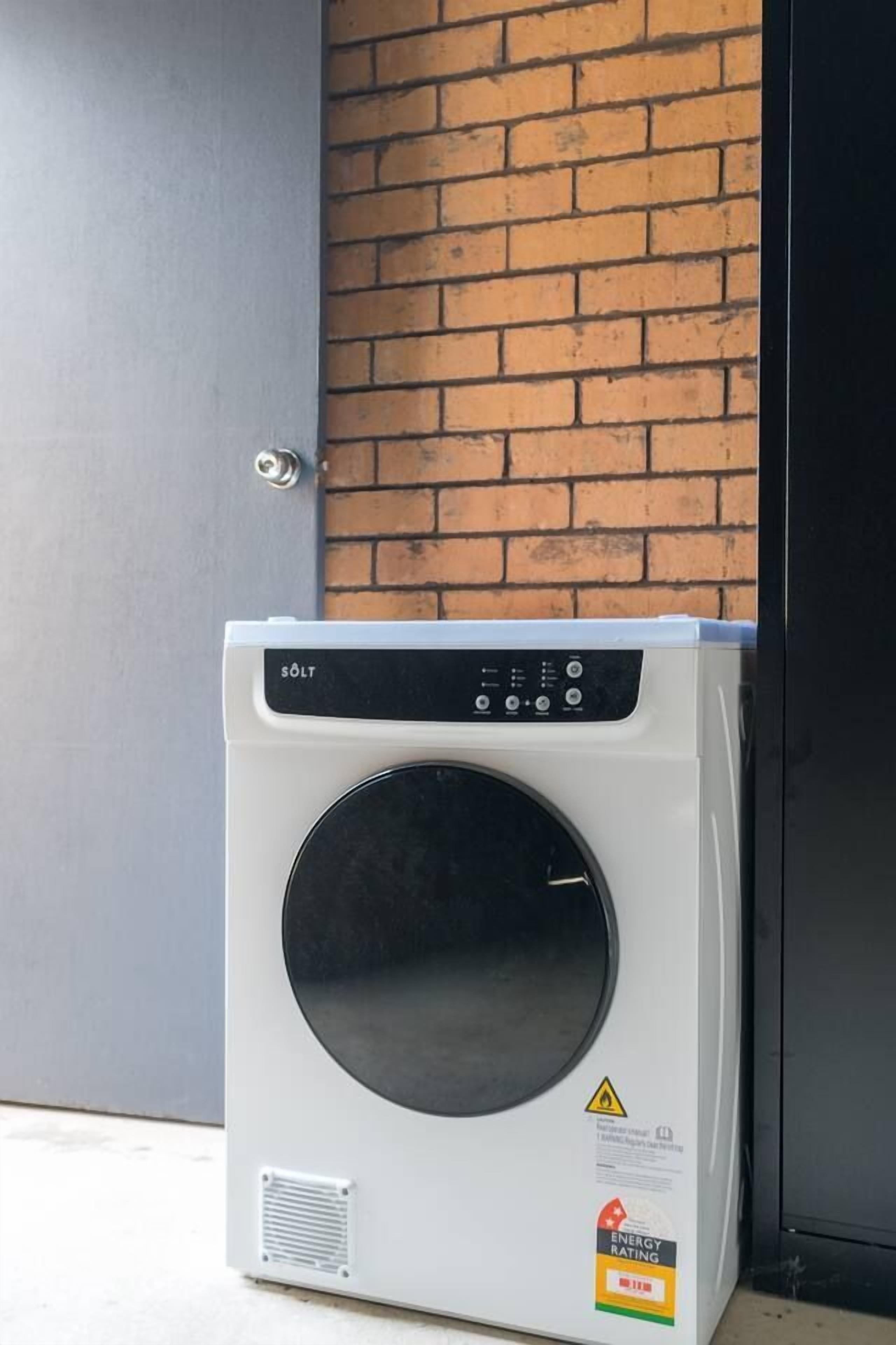 Une machine à laver blanche, dotée d’un panneau de commande, est placée contre un mur de briques, à côté d’une porte.