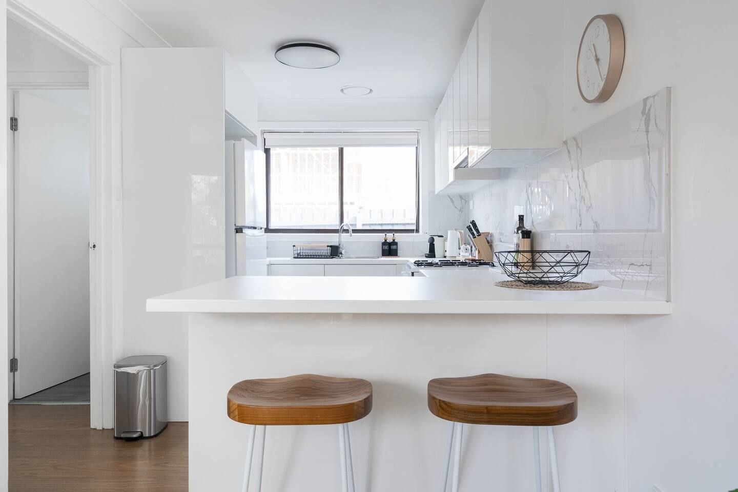 Een moderne keuken met een wit aanrecht, twee houten stoelen, een roestvrijstalen prullenbak en een raam met uitzicht.