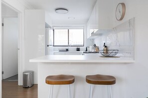 A modern kitchen with a white countertop, two wooden stools, a stainless steel trash can, and a window with a view. - Bayside Basking Coastal Rye Living at its Best (Rye)