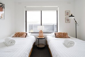 A hotel room with two beds, a window with blinds, a small table with a lamp, and a picture frame on the wall. - Bayside Basking Coastal Rye Living at its Best (Rye)