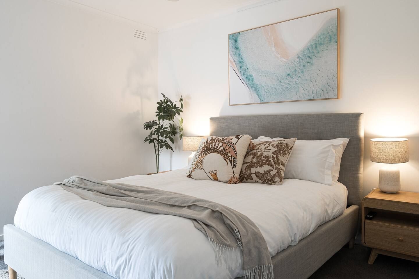 Un dormitorio con cama, lámparas de noche, una planta y un cuadro abstracto en la pared.
