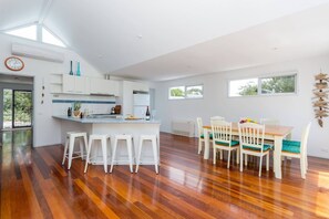 A modern kitchen with white cabinets, a central island with stools, and a dining area with wooden chairs and a table. - Stroll to the Sea Sun drenched Seaside Lifestyle (Rye)