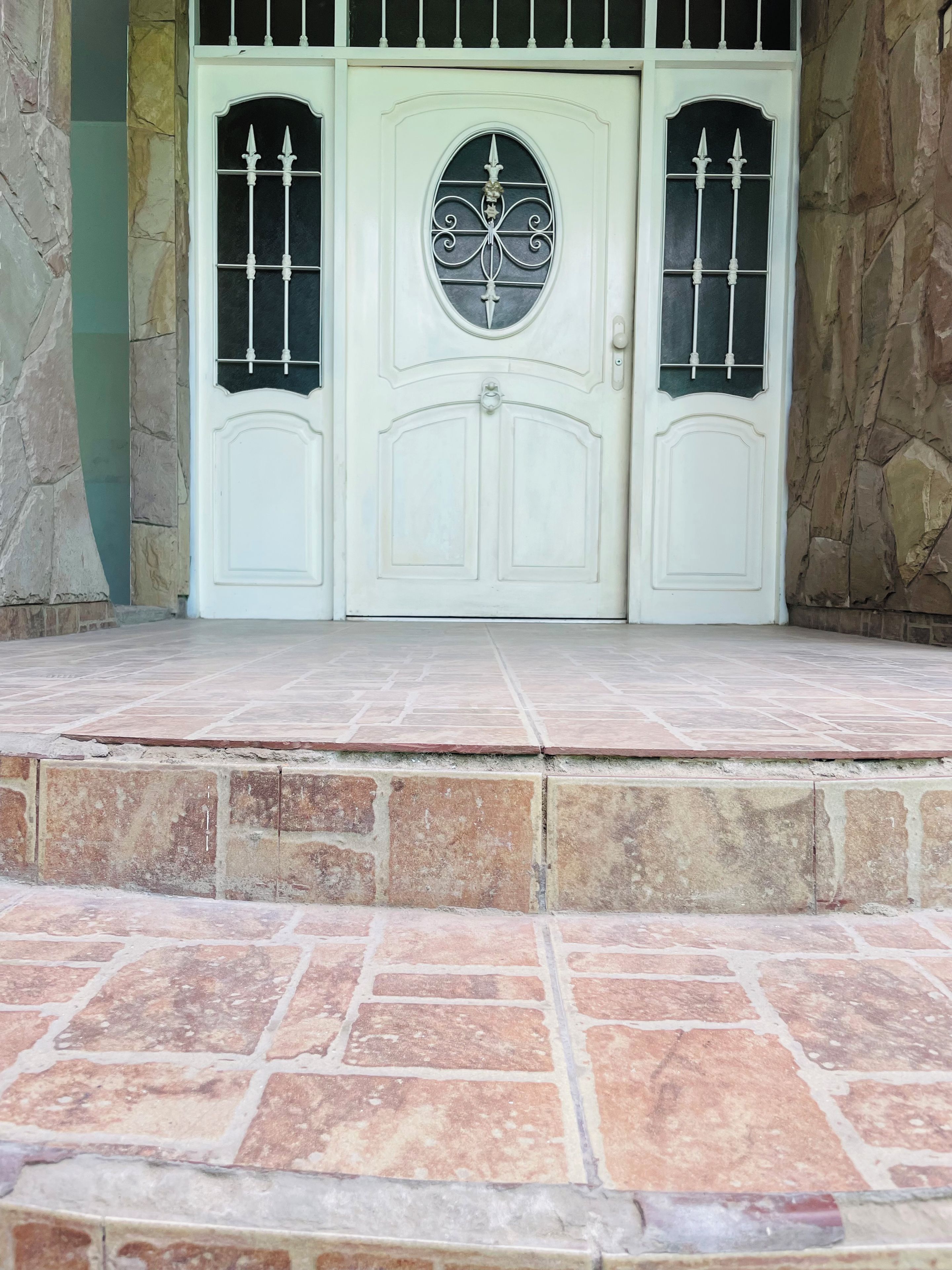 Uma porta dupla branca, com uma chapa decorativa em formato de olho de fechadura e uma grade de metal, emoldurada por uma parede de pedra.