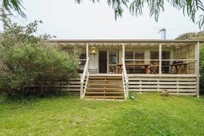 Ein eingeschossiges Haus mit überdachter Veranda, einer Holztreppe zum Eingang und einer kleinen Außenbereich-Sitzgruppe mit Tischen und Stühlen.