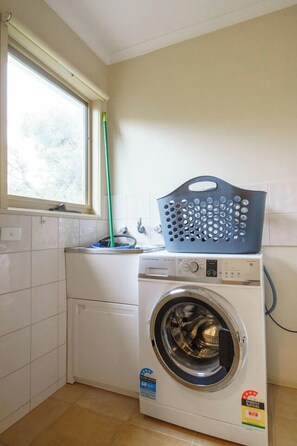 A compact laundry room with a front-loading washing machine, a drying rack, and a mop leaning against the wall. - Koonya Keeper Family Living by the Coast (Sorrento)