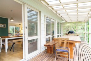 Eine überdachte Terrasse mit Holztisch und -stühlen, einem Grill und Blick ins Freie durch große Fenster.