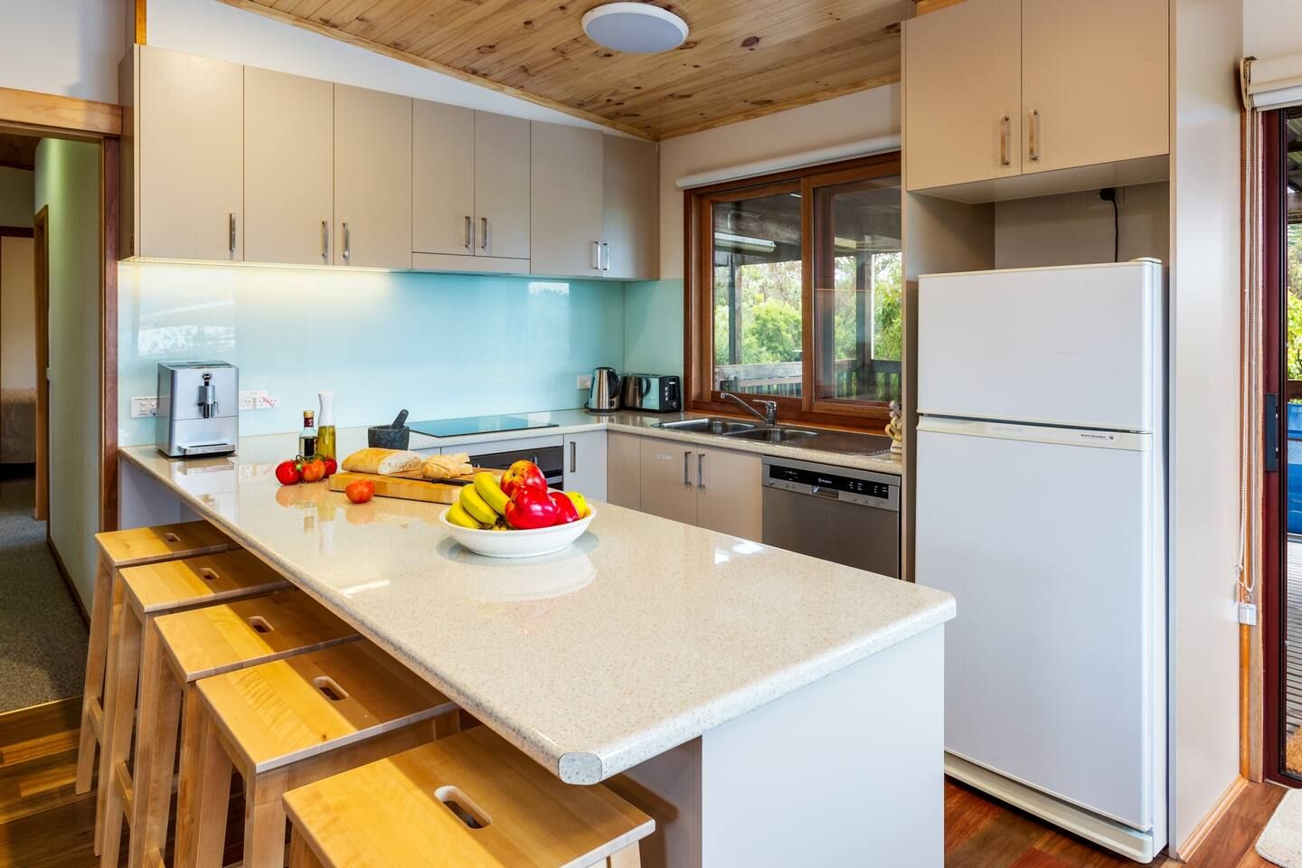 Une cuisine moderne dotée d’un îlot central, de tabourets en bois, d’un réfrigérateur et d’une fenêtre donnant sur un paysage verdoyant.