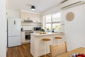 Eine kompakte Küche mit Kühlschrank, Herd, Backofen und Mikrowelle. Ein Fenster mit Jalousien, eine Uhr und ein kleiner Essbereich mit Barhocker.