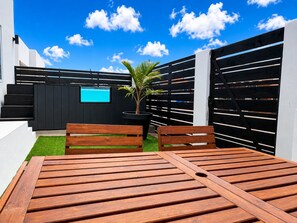 A patio with a wooden table and benches, a black fence, a palm tree, and a TV screen. - Brand NEW Calishi Apartment; Comfort & Style (Oranjestad)