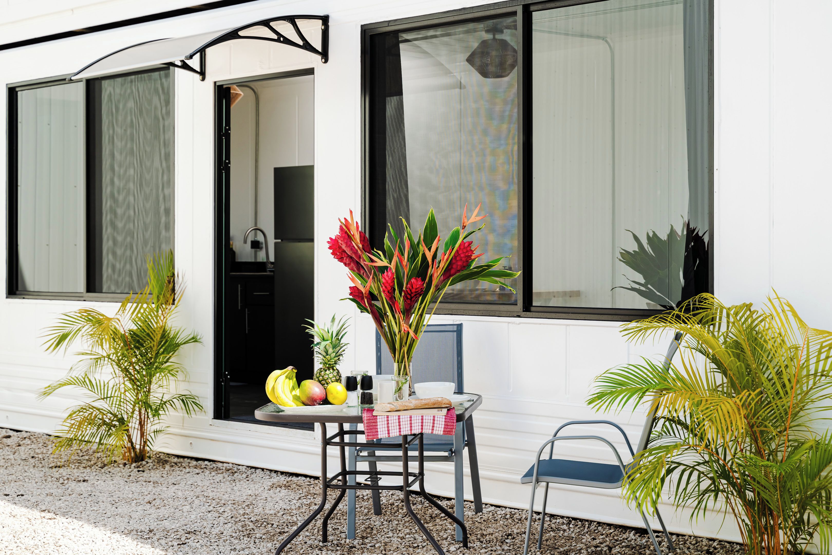 A white building with large windows, a table with a checkered tablecloth, a chair, and potted plants.