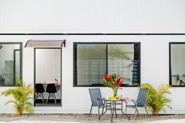 Un edificio moderno con ventanas amplias, un comedor con sillas y una mesa, y dos plantas en macetas.