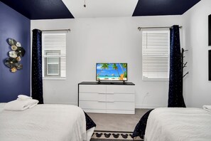 Ein Schlafzimmer mit zwei Betten, einem Schminktisch, einem Fernseher der eine Strandansicht zeigt und einem Fenster mit Blick auf Palmen.