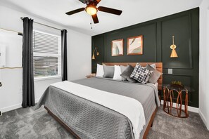 Quarto com cama, ventilador de teto, janela com persianas e pequena mesa com cadeiras.