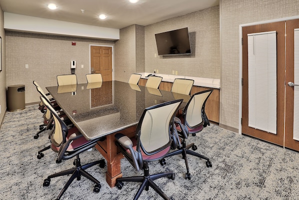 A conference room with a glass table surrounded by chairs, a mounted TV, and a door with blinds.