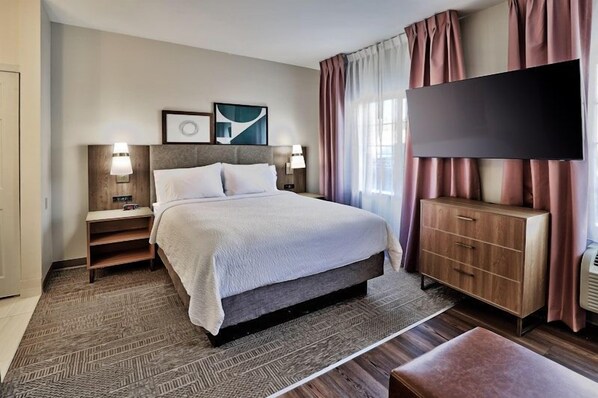 A hotel room with a large bed, a flat-screen TV, a wooden dresser, and a nightstand with a lamp.