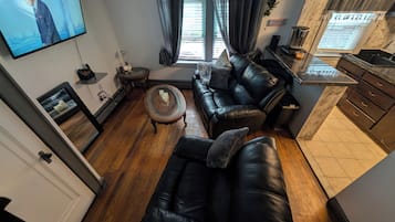 A living room with a black leather sofa, a coffee table, a TV mounted on the wall, a small side table, and a kitchen area with a countertop and cabinets.
