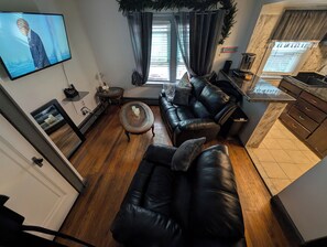 A living room with a black leather sofa, a coffee table, a TV mounted on the wall, a small side table, and a kitchen area with a countertop and cabinets.