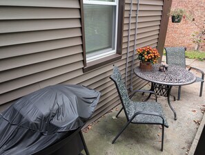 An outdoor seating area with a covered table and chairs, a window with blinds, and a brick wall.