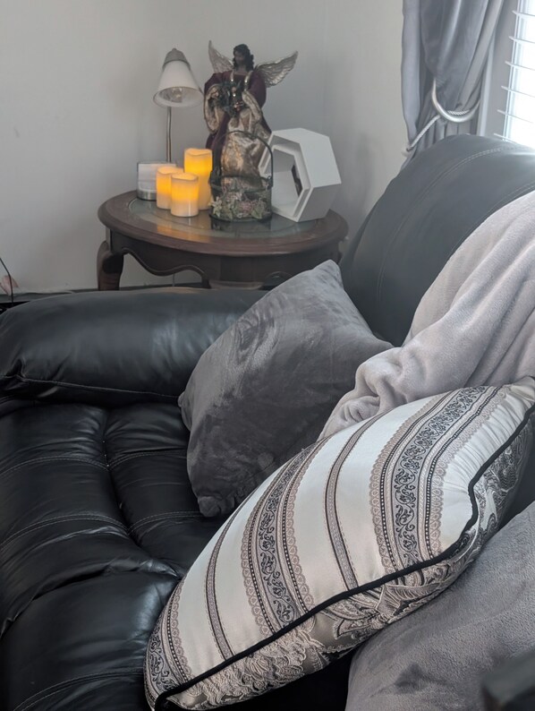 A black leather sofa with patterned cushions, a small table with a statue and candles, and a window with curtains.