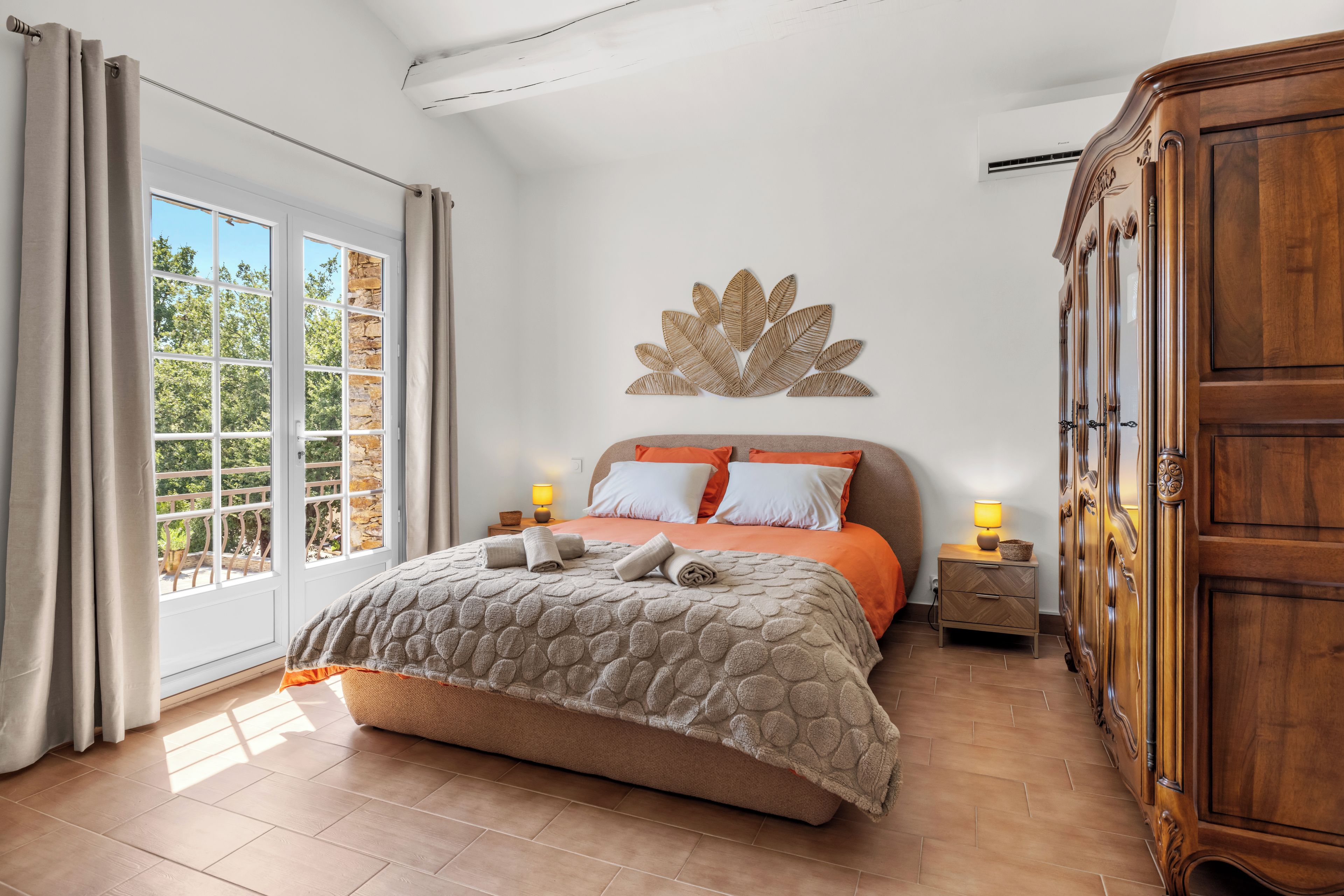 Un dormitorio con cama, mesita de noche, armario de madera y una ventana grande con cortinas.