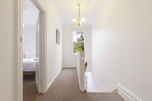 A hallway with a carpeted floor, a chandelier, a door leading to a bedroom, and a window with a view of greenery. - Skylight Terrace Timeless Stay near Parks and Cafs (Fitzroy North)
