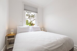 A bedroom with a large bed, two bedside tables, a window with a view of trees, and a wall-mounted light. - Skylight Terrace Timeless Stay near Parks and Cafs (Fitzroy North)