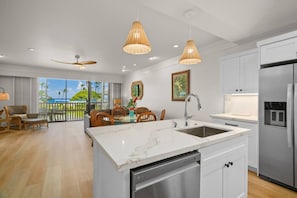 A modern kitchen with a large island, stainless steel appliances, and a dining area with a view. - Kiahuna Hale Kai II (Koloa)