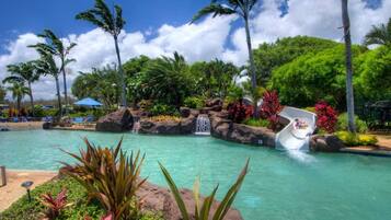 A pool with a water slide, surrounded by tropical vegetation and palm trees.
