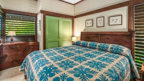 A bedroom with a bed, a nightstand, a lamp, a window with blinds, and a green door. - Kiahuna Hale Kai III (Koloa)
