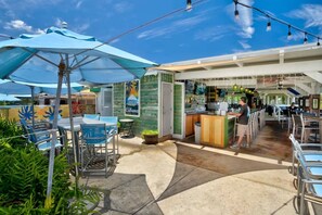 Uma área de refeições ao ar livre com mesas e cadeiras, um bar com balcão e uma parede verde com uma placa.