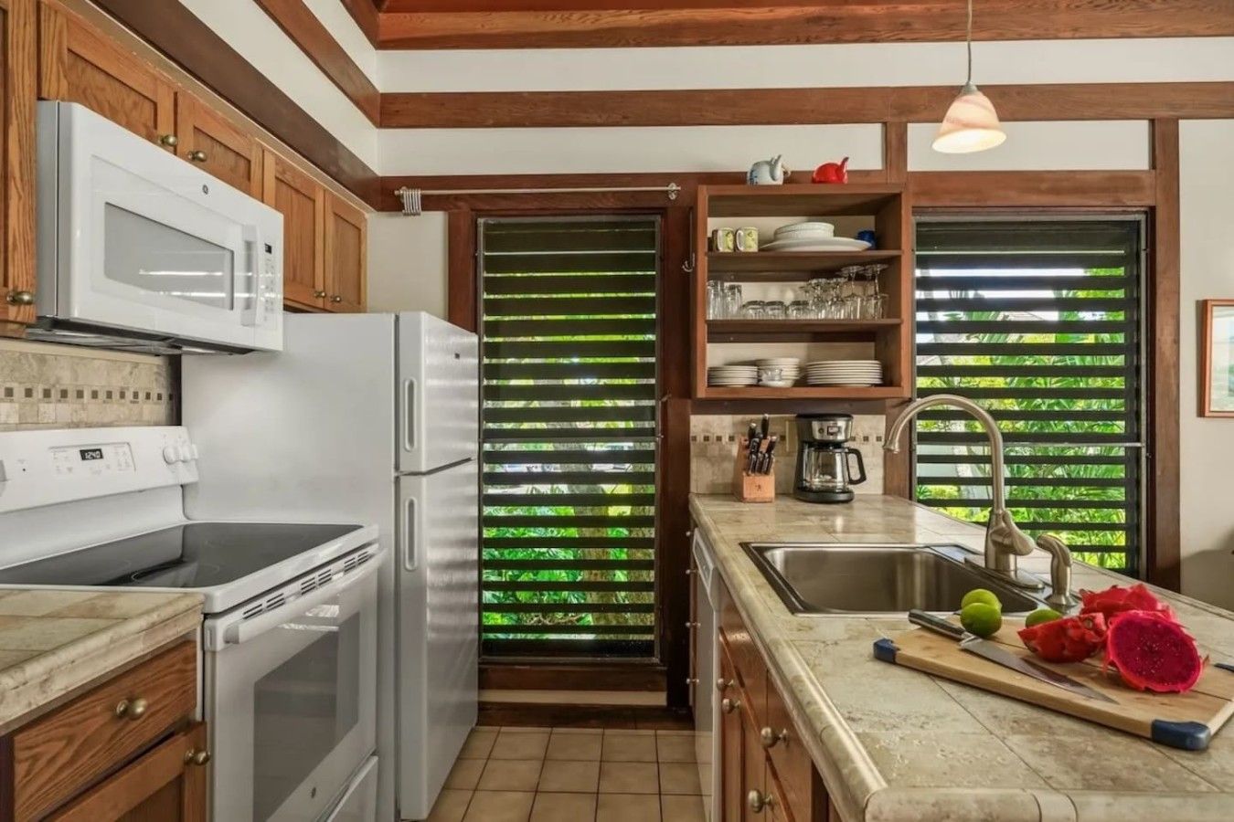 A kitchen with a refrigerator, microwave, stove, and sink. There are wooden cabinets and a window with green blinds.