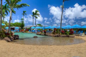 Un espace piscine aménagé dans un complexe hôtelier, avec des chaises longues et des parasols, entouré de palmiers et surplombé par un ciel dégagé.